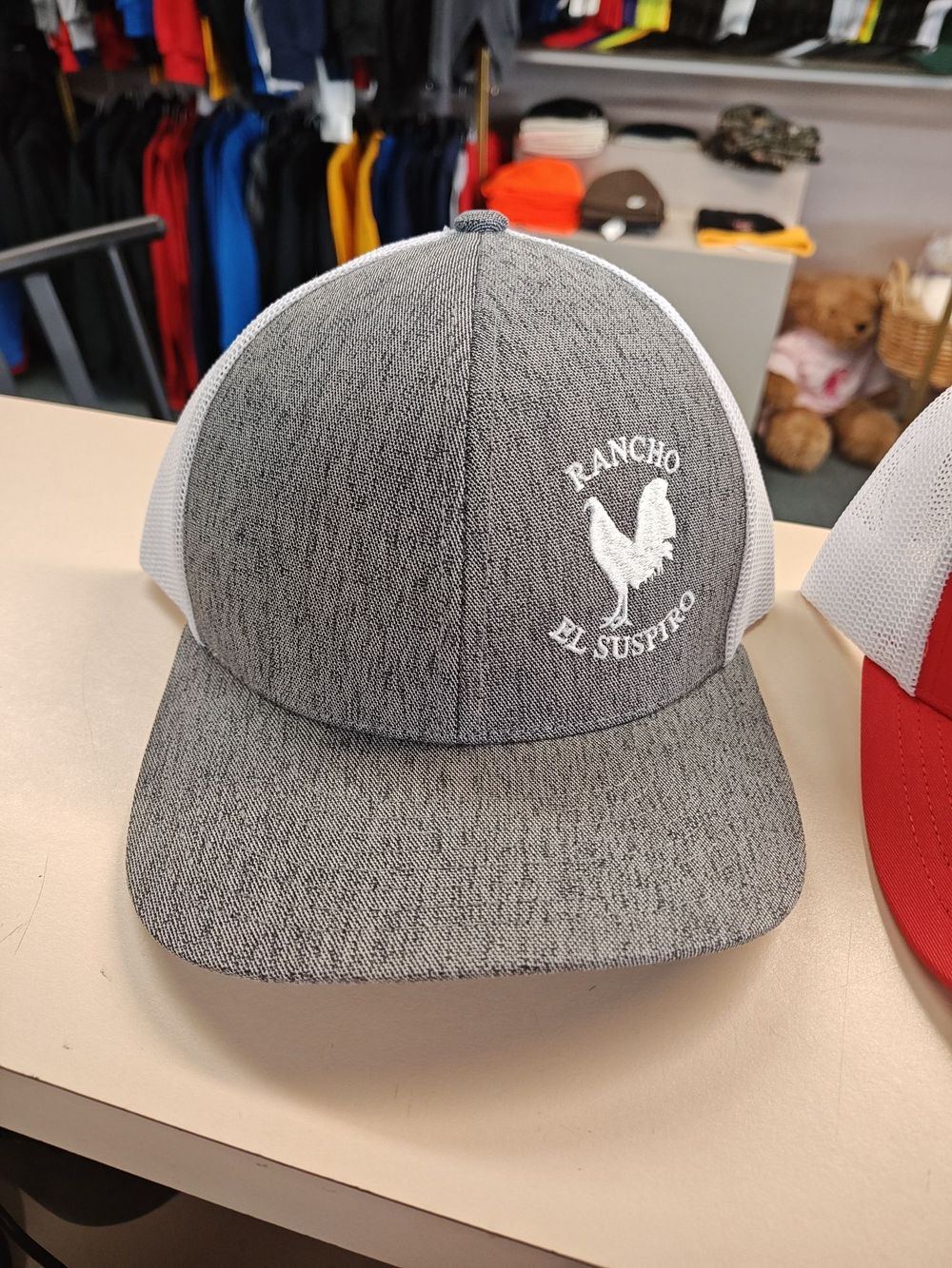 Grey trucker cap with a round white embroidered rooster logo reading Rancho El Suspiro