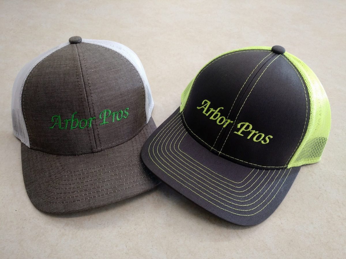 Two trucker caps, one brown and one charcoal with neon mesh, embroidered in green with Arbor Pros