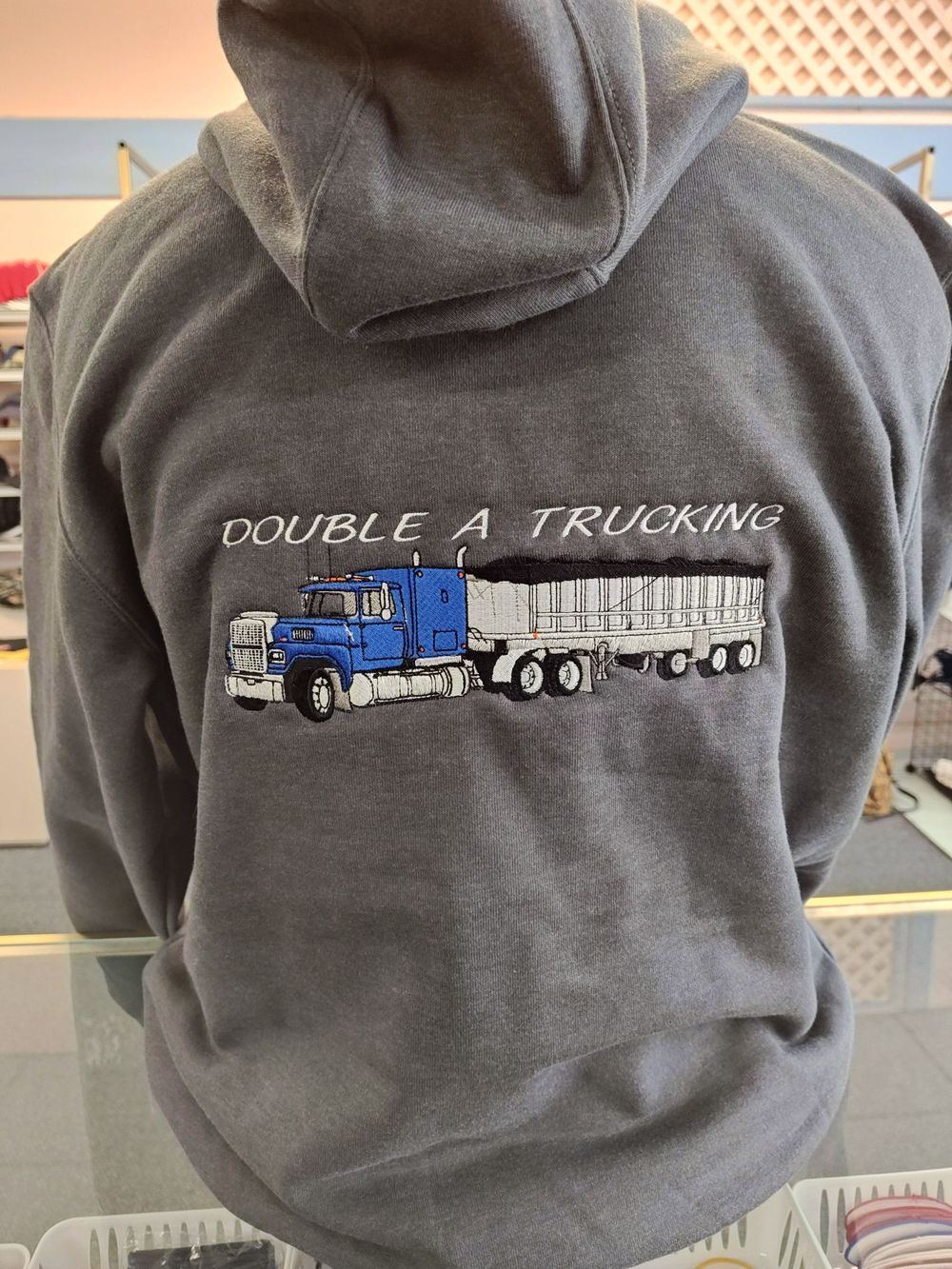 Back of a light grey hooded sweatshirt embroidered with a Double A Trucking logo and a blue semi-truck