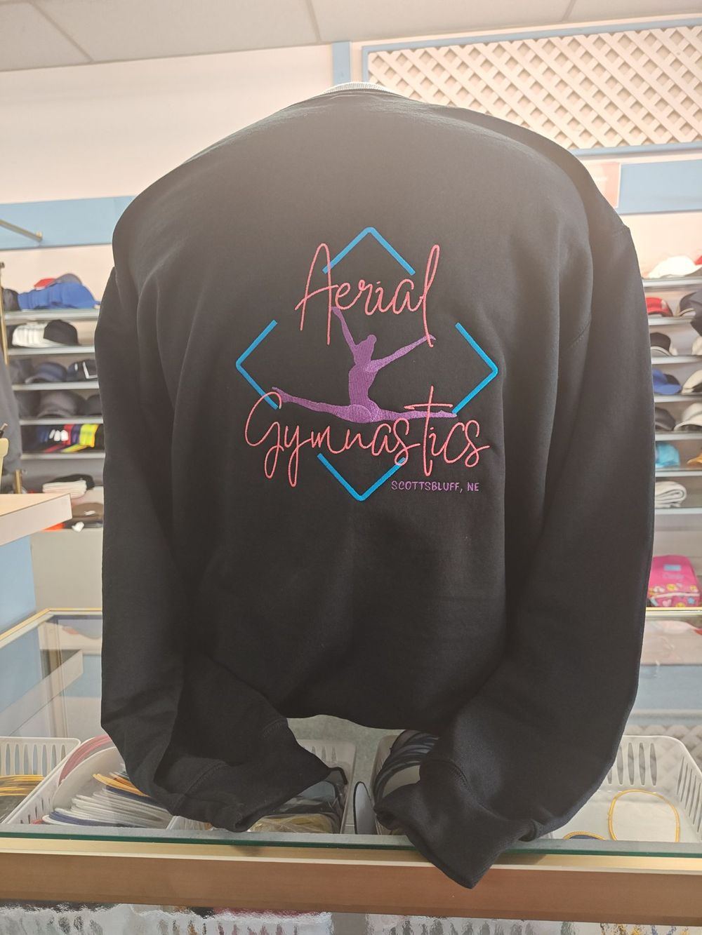 Back of a black sweatshirt with a colorful embroidered Aerial Gymnastics logo of a gymnast in a split jump